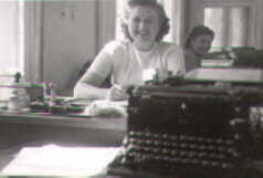 Schwarzweißfoto: im Vordergrund eine alte Schreibmaschine, im Hintergrund: eine Sekretärin.